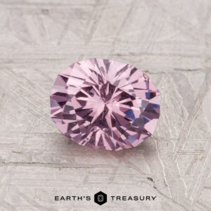 A round, faceted 1.34-carat garnet gemstone in light purple rests on a textured gray surface with the Earth's Treasury logo and text at the bottom.