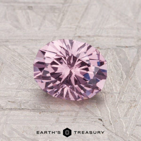 1.34-Carat Garnet A round, faceted 1.34-carat garnet gemstone in light purple rests on a textured gray surface with the Earth's Treasury logo and text at the bottom.