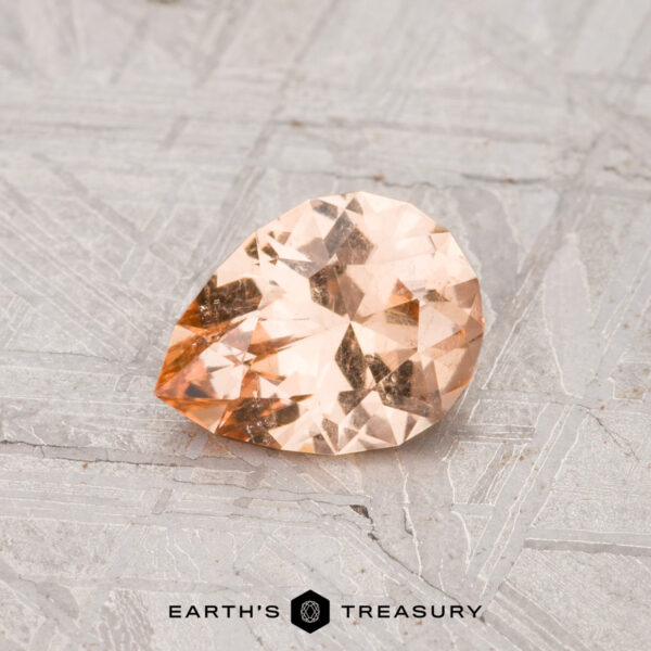 0.77-Carat Grossular Garnet A faceted, pear-shaped 0.77-carat grossular garnet with a peach hue rests on a textured gray surface, with the "EARTH'S TREASURY" text and logo displayed at the bottom.