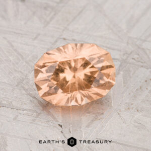 A 1.34-carat Grossular Garnet, faceted in an oval peach hue, displayed on a textured metallic surface with the Earth’s Treasury logo at the bottom.