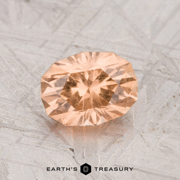 1.34-Carat Grossular Garnet A 1.34-carat Grossular Garnet, faceted in an oval peach hue, displayed on a textured metallic surface with the Earth’s Treasury logo at the bottom.