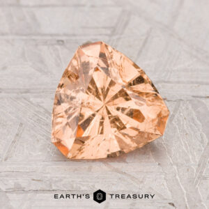 A 1.53-carat Grossular Garnet, cut in a trillion shape and peach-colored, is displayed on a textured gray surface with "Earth's Treasury" shown at the bottom.