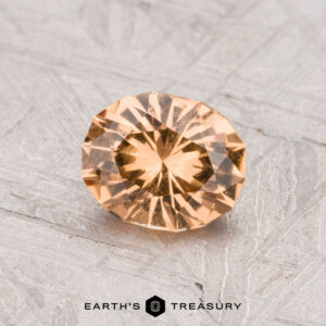 A 0.97-carat round, faceted Grossular Garnet with a peach hue is displayed on a gray, patterned surface. "EARTH'S TREASURY" logo and text are visible at the bottom of the image.
