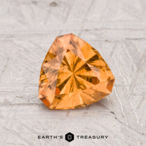 A faceted, triangular 1.26-carat Grossular Garnet with an orange hue is displayed on a textured light gray surface, featuring the "Earth's Treasury" logo and text at the bottom.