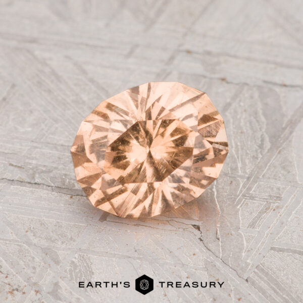 1.21-Carat Grossular Garnet A 1.21-carat round, faceted grossular garnet with a peach hue rests on a textured gray surface, featuring the text "EARTH'S TREASURY" at the bottom.
