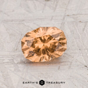 A 1.17-carat grossular garnet, oval and faceted with an orange hue, rests on a textured metallic surface displaying "EARTH'S TREASURY" at the bottom.
