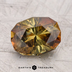 A 3.42-carat, faceted oval Andalusite with yellow and brown hues rests on a textured gray surface. The "EARTH'S TREASURY" logo appears at the bottom.
