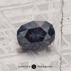 A 1.42-carat color change garnet rests on a textured metallic surface, with "EARTH'S TREASURY" displayed at the bottom.