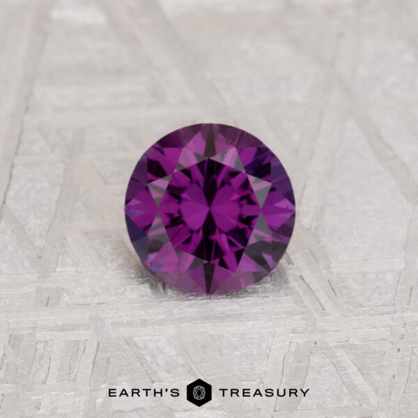 0.56-Carat Color Change Garnet A round, faceted 0.56-carat Color Change Garnet gemstone displayed on a light textured surface with the logo and text "EARTH'S TREASURY" at the bottom.