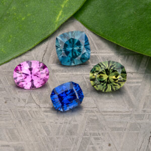 Stacked from 16 images. Method=B (R=9,S=5), four faceted gemstones in pink, blue, green, and teal rest on a patterned surface beside two green leaves.