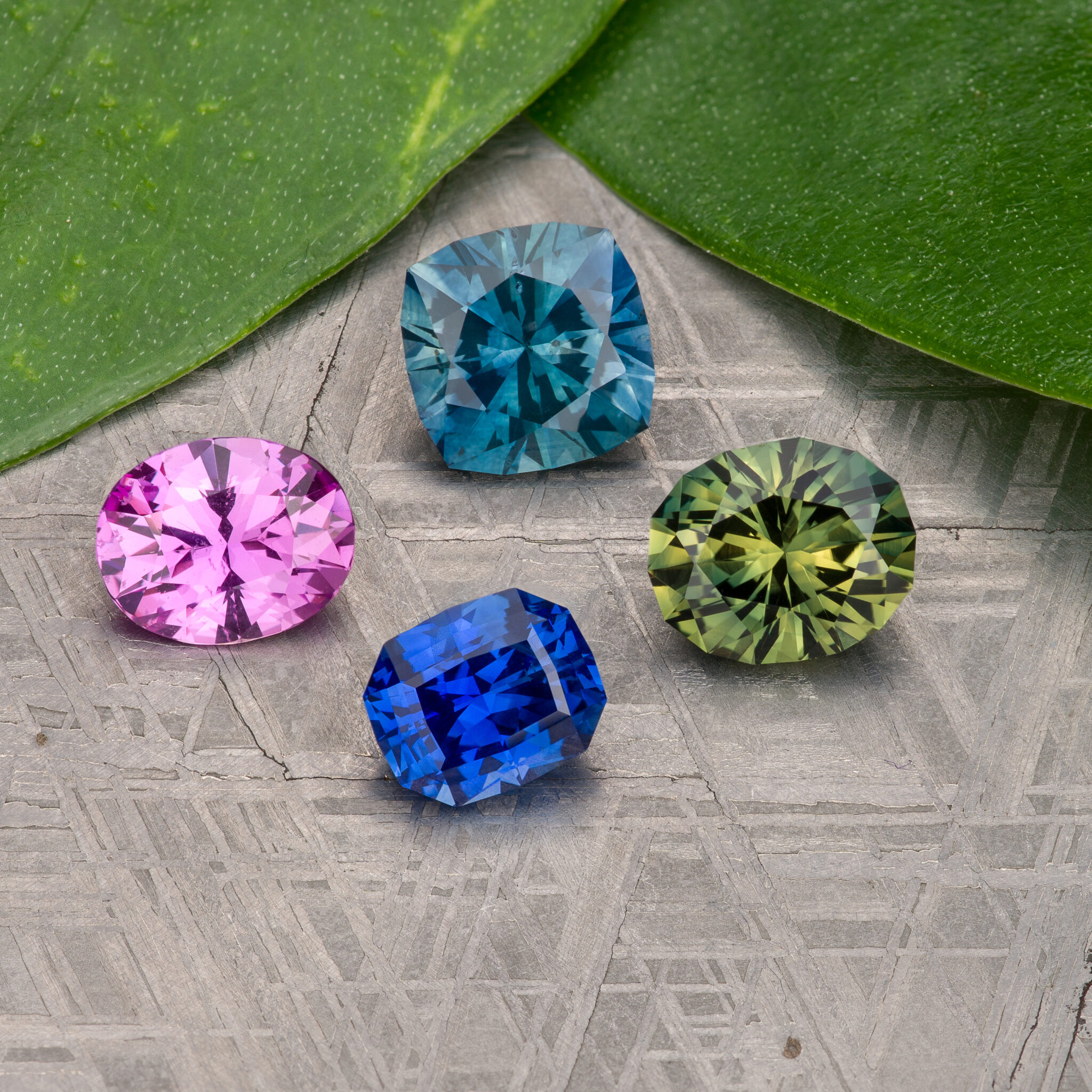 Stacked from 16 images. Method=B (R=9,S=5), four faceted gemstones in pink, blue, green, and teal rest on a patterned surface beside two green leaves.