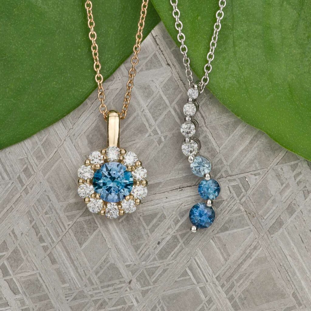 Two all-jewelry necklaces featuring blue gemstones and diamonds are showcased on a silver textured surface with green leaves in the background.