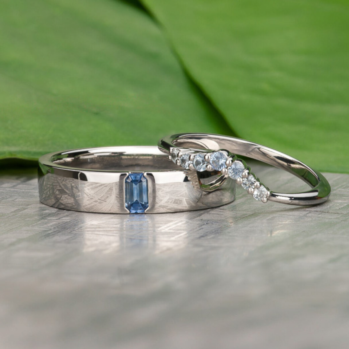 Two silver bands from Earth's Treasury lie on a textured surface—one set with a blue rectangular gem, the other featuring a curved row of small clear stones. Green leaves provide a subtle background.