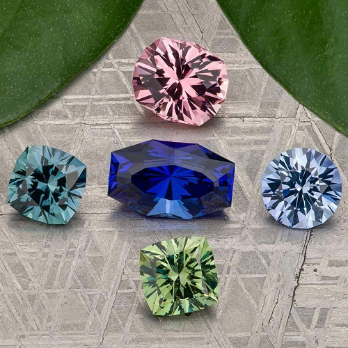 Five faceted gemstones in blue, pink, green, and light blue hues from "choosing-a-colored-gemstone" are displayed on a patterned gray surface, with two green leaves partially visible at the top.