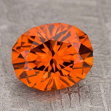 The gem-color-orange, a faceted oval gemstone, is shown on a textured gray surface.
