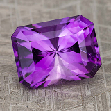 A rectangular, faceted gemstone named gem-color-purple is displayed on a textured gray surface.