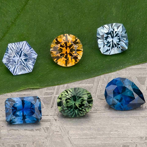 Six faceted gemstones in varied shapes and colors—blue, yellow, and green—from the gem-size-guide are displayed on a green leaf and a gray textured surface.