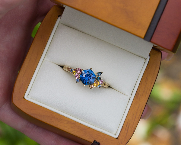 The propose ring features a large blue gemstone accented by smaller multicolored stones, showcased in an open wooden box with a white cushioned interior.