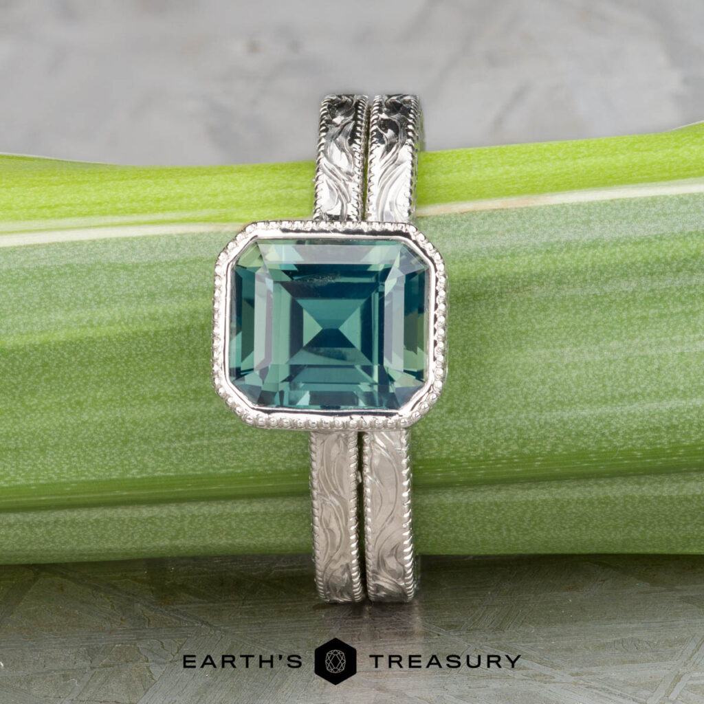 A silver Straight Astrea Band by Johnson, Dalton and Murphy, Erin with an emerald-cut green gemstone is displayed on a green stem, with "Earth's Treasury" logo and text below.