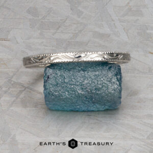 The ET Stock silver textured Straight Astrea Band is displayed on a rough blue gemstone atop a gray patterned surface, with the Earth’s Treasury logo beneath.