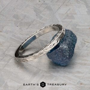A silver ET Stock band with engraved patterns rests on a rough blue stone against a textured gray background. The text "EARTH'S TREASURY" appears below.