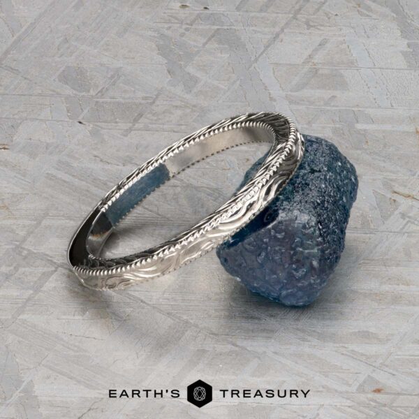 The Custom-Fit Engraved band in platinum A silver ET Stock band with engraved patterns rests on a rough blue stone against a textured gray background. The text "EARTH'S TREASURY" appears below.