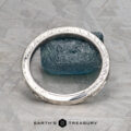A silver ET Stock bangle with intricate patterns stands upright, encircling a rough blue-green stone on a gray textured surface. The phrase “Earth’s Treasury” is displayed below.