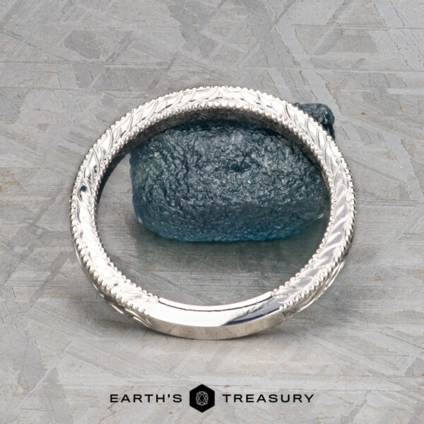 The Custom-Fit Engraved band in platinum A silver ET Stock bangle with intricate patterns stands upright, encircling a rough blue-green stone on a gray textured surface. The phrase “Earth’s Treasury” is displayed below.