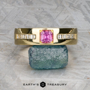 A Custom ring in 18k royal yellow gold with a 0.42-carat Montana sapphire is displayed on a rough blue-green mineral surface, with "Earth's Treasury" text below.