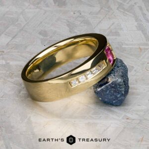 A custom ring in 18k royal yellow gold featuring a 0.42-carat Montana sapphire rests on a rough dark mineral atop a textured gray surface.