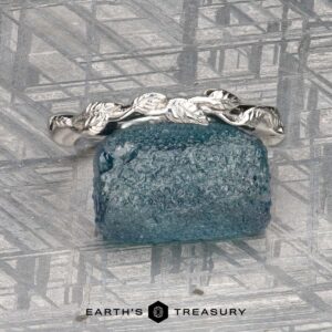 The Mooney, Mia and Wight, Spencer 2 ring—a silver band with a leaf motif inspired by Guinevere—rests on a rough blue gemstone, set before a metallic background with "Earth's Treasury" displayed below.