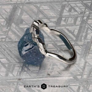 A silver Mooney, Mia and Wight, Spencer 2 ring with a rough blue gemstone is displayed on a metallic, patterned background. The text "EARTH'S TREASURY" appears at the bottom.
