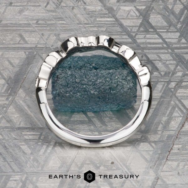The "Linden" band in platinum The Mooney, Mia and Wight, Spencer 2 ring—a silver band with a rough blue gemstone—rests on a geometric-patterned surface above the “Earth’s Treasury” text, capturing Guinevere-like elegance.