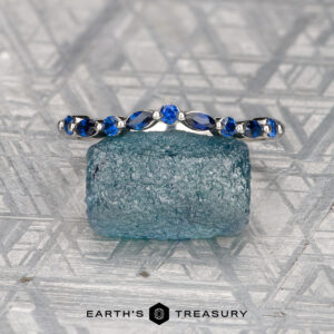 A Mock, Eli and Herbert, Olivia 2 ring—silver with oval and round sapphires—rests on a rough blue crystal atop a geometric patterned background, with "EARTH'S TREASURY" beneath.