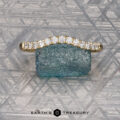 An Obendorf, Kenny and Kitchin, Amelia 2 gold ring with small diamonds is shown resting on a rough blue-green stone against a patterned metallic backdrop. The text "EARTH'S TREASURY" appears below.