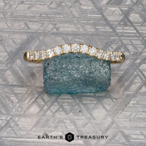 An Obendorf, Kenny and Kitchin, Amelia 2 gold ring with small diamonds is shown resting on a rough blue-green stone against a patterned metallic backdrop. The text "EARTH'S TREASURY" appears below.