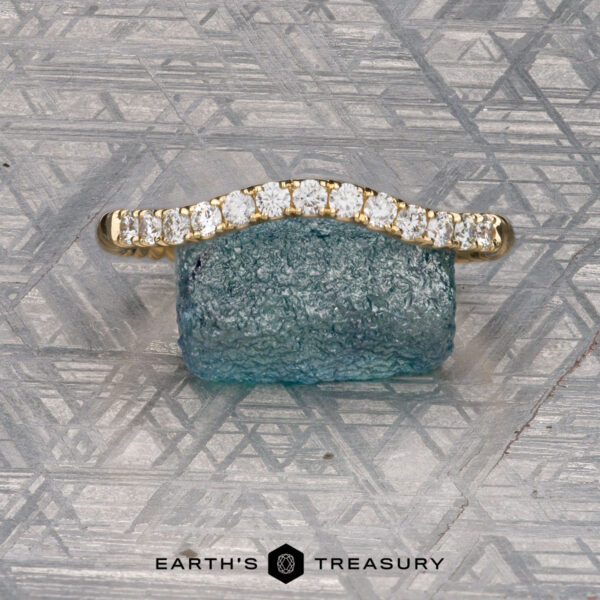 The Contour "Lynn" band in 18k yellow gold An Obendorf, Kenny and Kitchin, Amelia 2 gold ring with small diamonds is shown resting on a rough blue-green stone against a patterned metallic backdrop. The text "EARTH'S TREASURY" appears below.