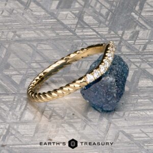 The Obendorf, Kenny and Kitchin, Amelia 2 ring, a gold twisted band with small diamonds, sits atop a rough blue gemstone on a geometric metallic surface. The "Earth's Treasury" logo appears at the bottom.