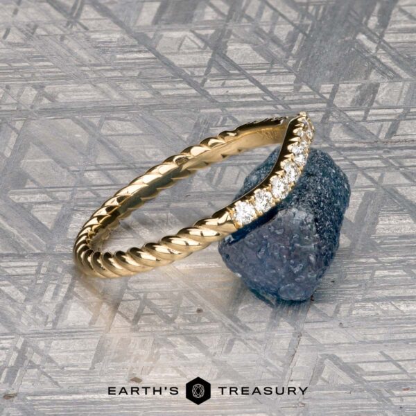 The Contour "Lynn" band in 18k yellow gold The Obendorf, Kenny and Kitchin, Amelia 2 ring, a gold twisted band with small diamonds, sits atop a rough blue gemstone on a geometric metallic surface. The "Earth's Treasury" logo appears at the bottom.