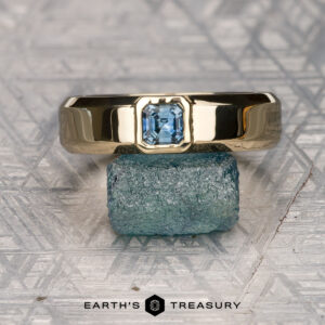 The product "Stacked from 16 images. Method=B (R=10,S=7)" is showcased as a classic gold Sapphire Ring with a square-cut blue gemstone, standing on a rough blue stone atop a patterned silver surface marked "EARTH'S TREASURY.