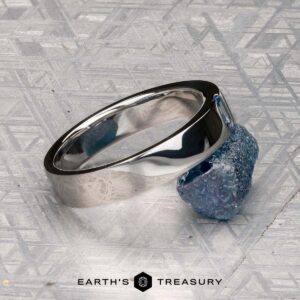 The Harrison, Samuel 3 ring, a polished silver classic sapphire design, rests on a rough blue stone atop a textured metallic surface, with the "Earth's Treasury" logo featured at the bottom.