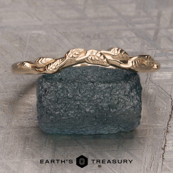 The "Linden" band in 14K yellow gold The "Linden" band in 14K yellow gold, featuring a delicate leaf design, is displayed atop a rough blue gemstone with the text "EARTH'S TREASURY" below.