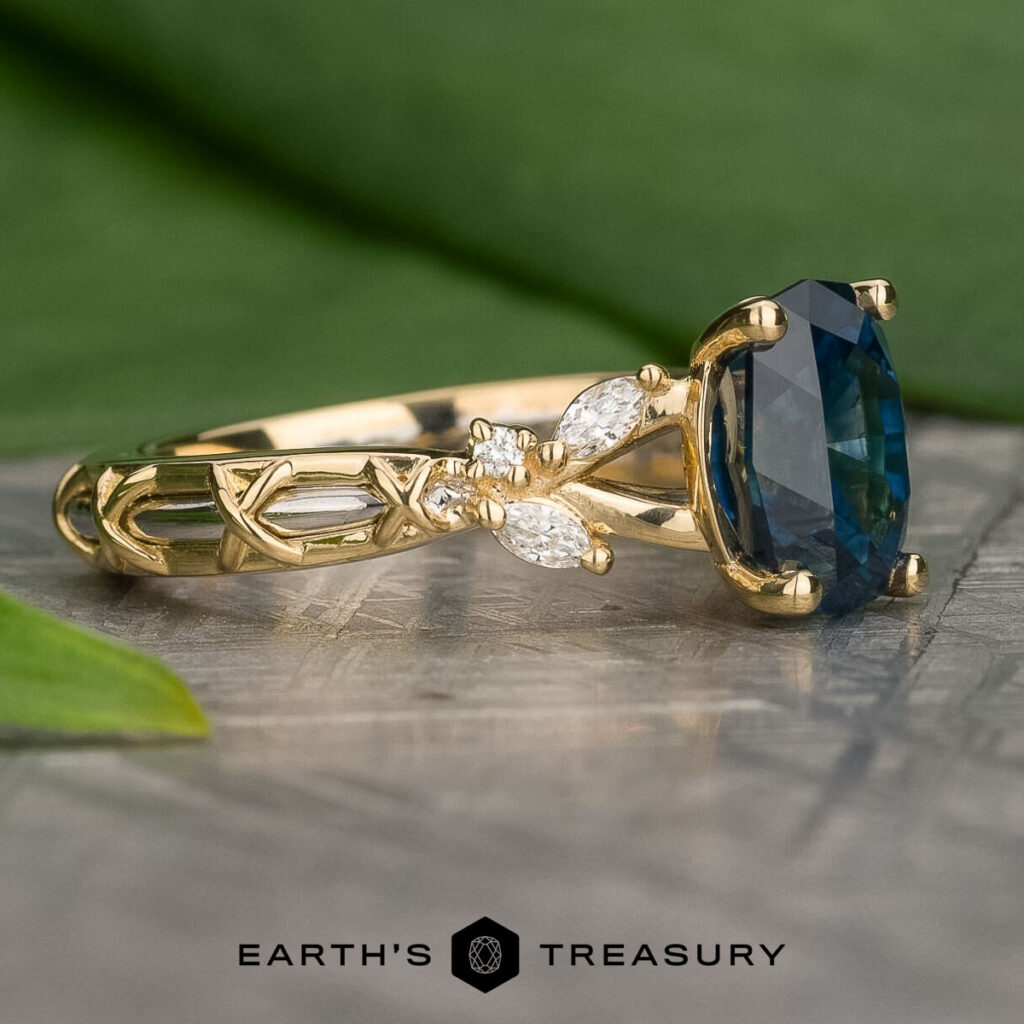 Product 4f9679f7-de93-46ce-857c-bf260fcf8300-1200-BW, a gold ring with an oval blue gemstone and two marquise-cut clear stones on a detailed band, is displayed on a stone surface.