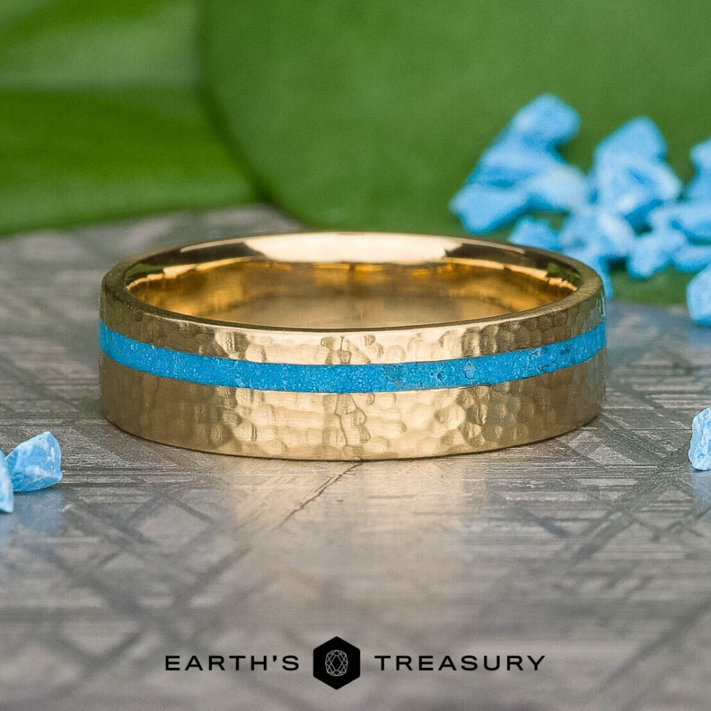 The 5b0b7134-183d-4748-96c7-f23ed76c03ad-1200-BW gold ring with a hammered texture and blue inlay rests on a patterned surface, surrounded by blue crystals and green leaves. The text reads: "EARTH'S TREASURY.