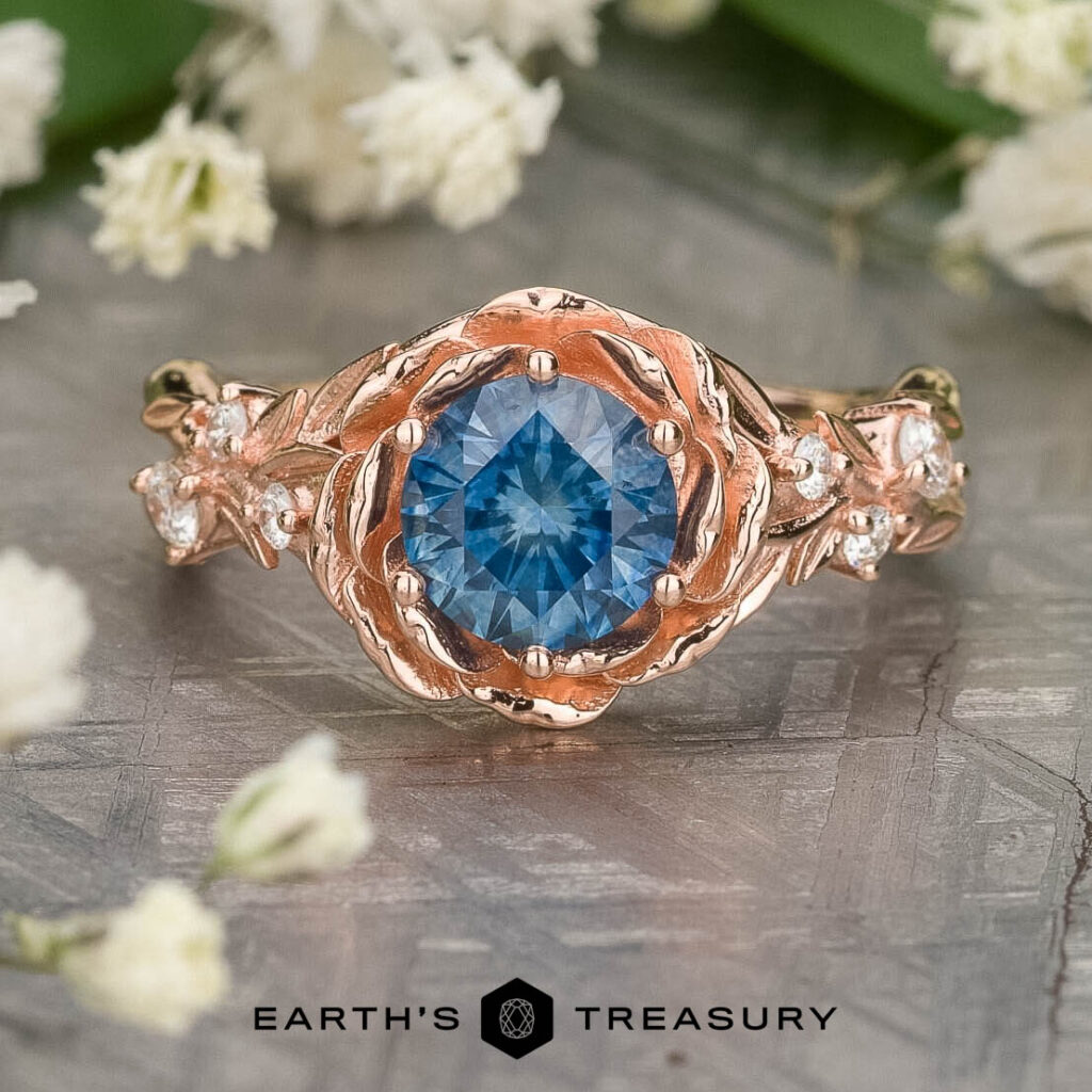 The 7babe330-10f4-4dd6-9872-ae46da9e9725-1200-BW-2 ring features a rose gold band with a round blue center stone, leaf accents, and white stones, shown on a textured surface beside white flowers.