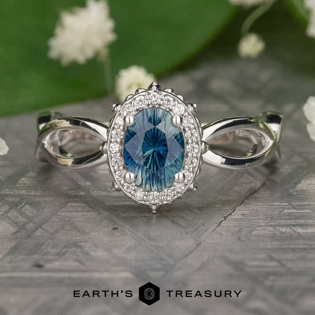 Product 91204c13-c0d2-4374-9a31-f02514baad19-1200-BW: A silver ring with an oval blue gemstone, clear accent stones, and an infinity band, shown on a textured surface with green leaves and flowers in the background.