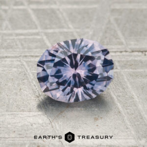 A round, faceted 1.37-carat heated Montana Sapphire sits on a textured gray surface with the "EARTH'S TREASURY" logo beneath it.