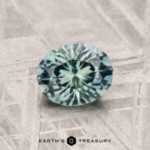 A 1.12-carat round, faceted blue-green Montana Sapphire gemstone is shown on a textured, patterned surface with "EARTH'S TREASURY" at the bottom.