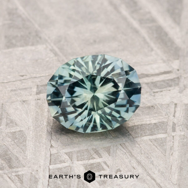 1.12-Carat Montana Sapphire A 1.12-carat round, faceted blue-green Montana Sapphire gemstone is shown on a textured, patterned surface with "EARTH'S TREASURY" at the bottom.
