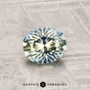 A 1.01-carat Montana Sapphire with blue and yellow hues, faceted in an oval cut, rests on a textured gray surface. "EARTH'S TREASURY" is displayed at the bottom of the image.
